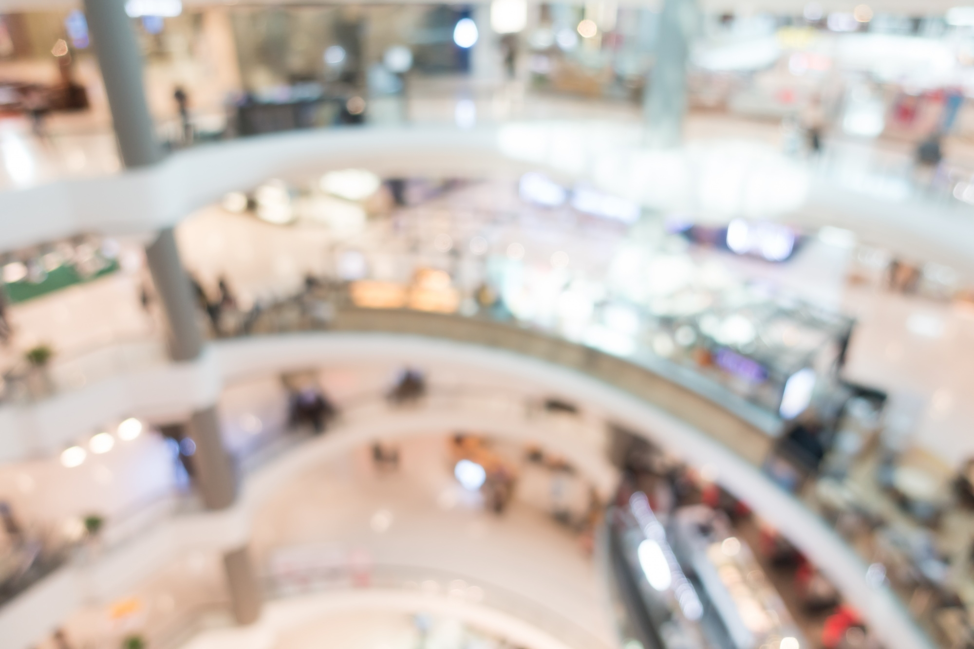 Assosia 5 Blurred interior of a bright shopping mall with an escalator, storefronts, and shoppers walking through the space.