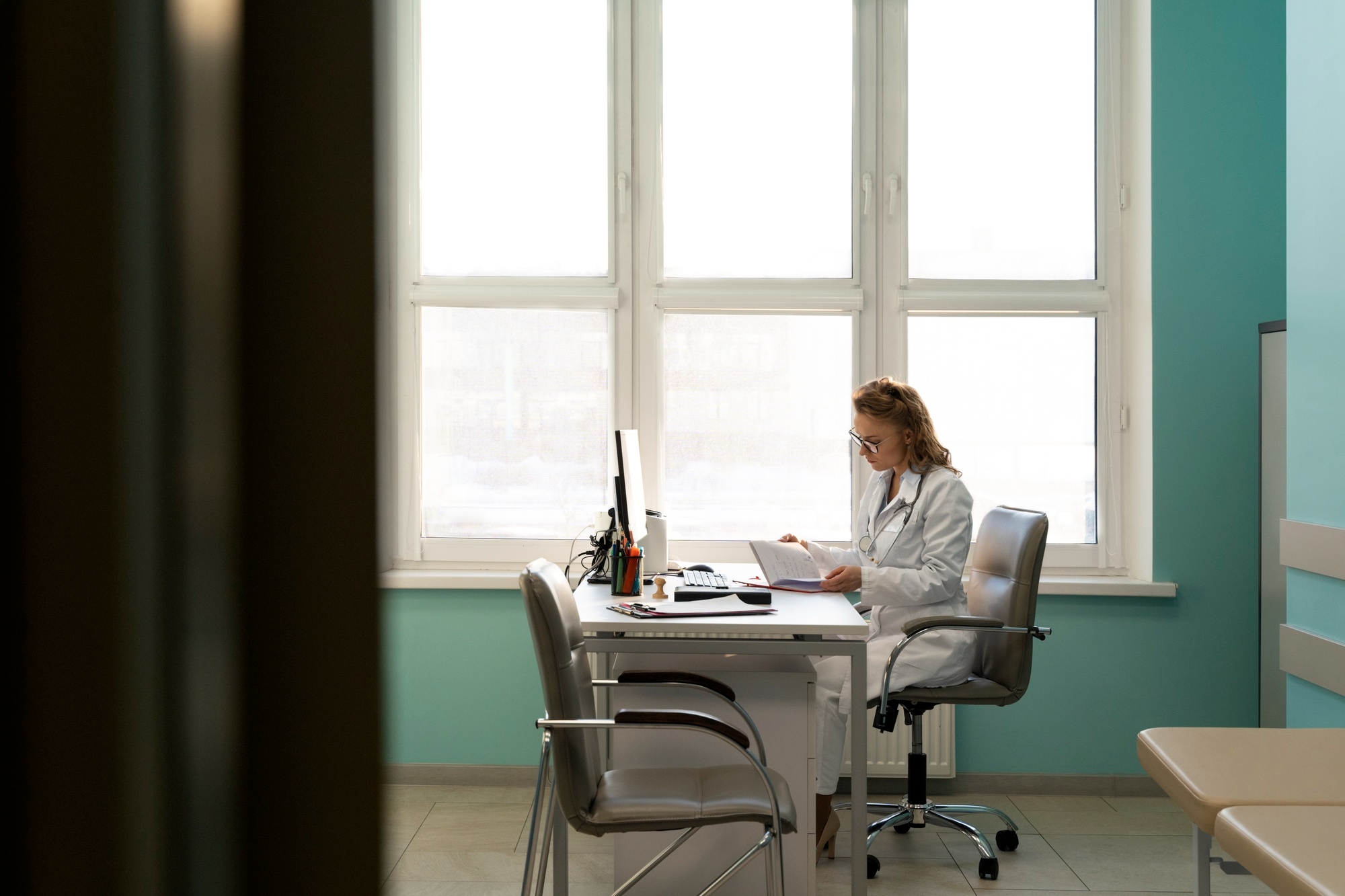 Female doctor in a white coat and glasses sitting at a desk reviewing a document, with a computer monitor and teal-painted walls in the background.