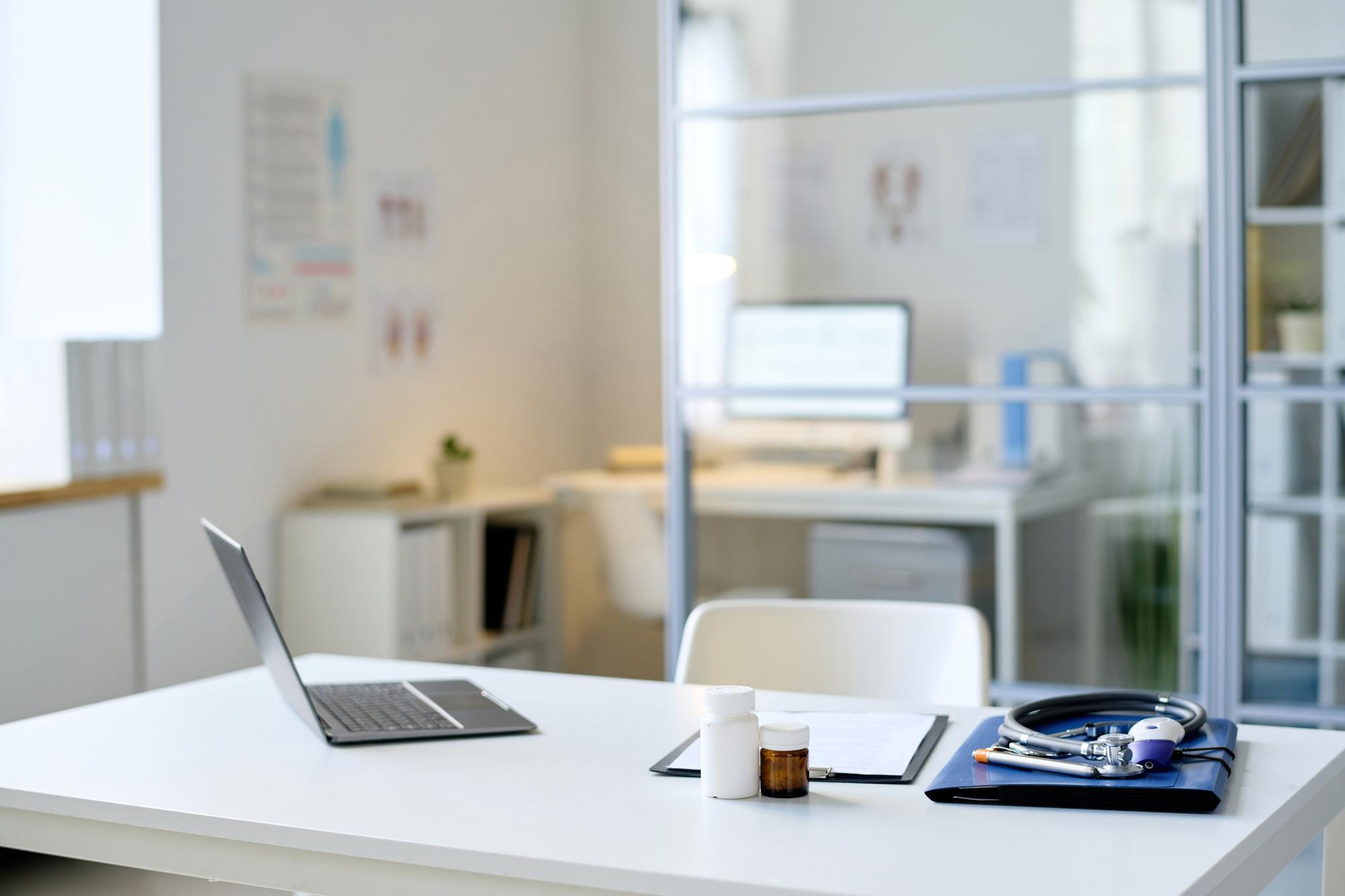 Empty doctor's office desk with an open laptop, two medicine bottles, a clipboard, and a stethoscope on a blue case, with a second workstation visible in the background.