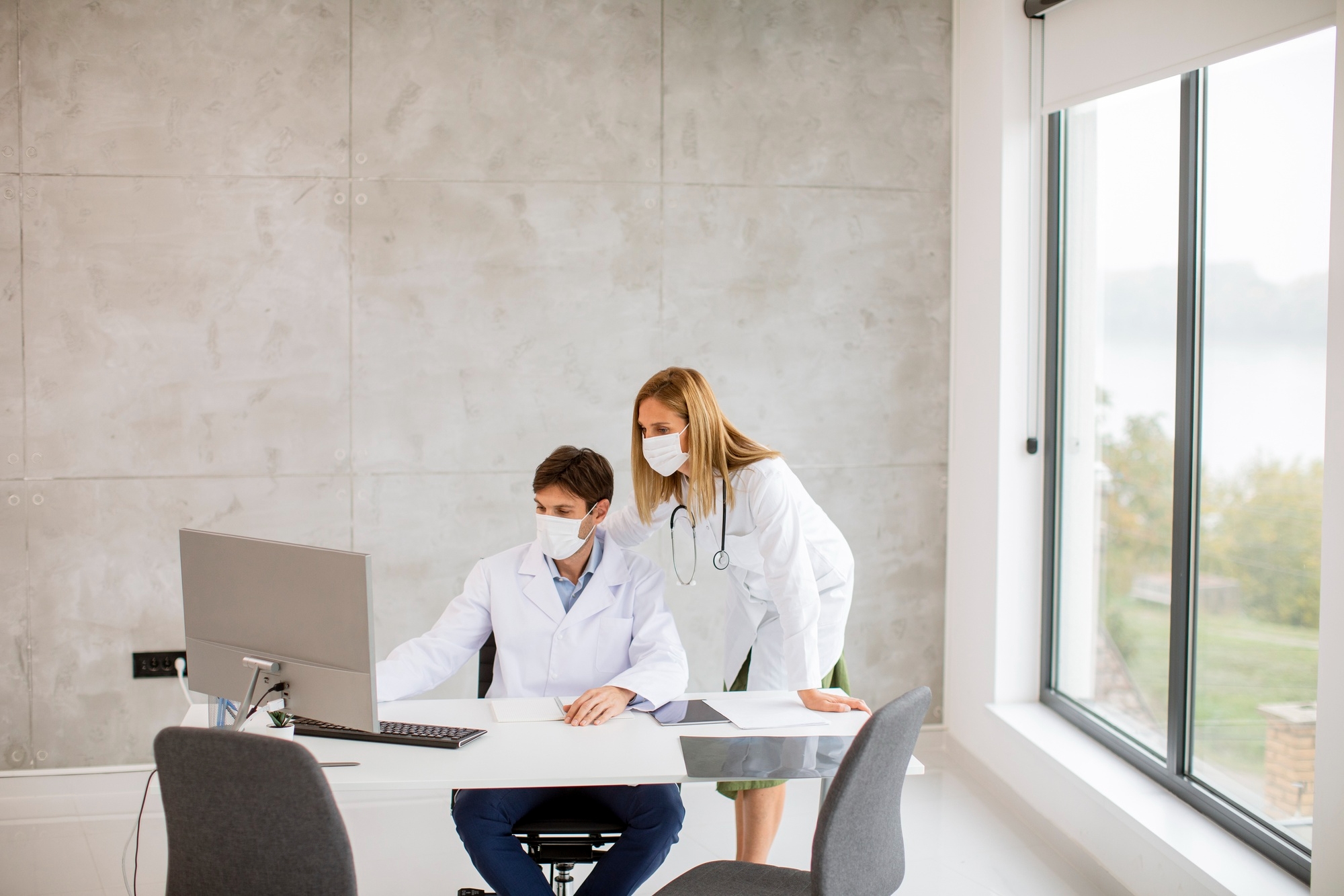 Two medical professionals in white coats and face masks reviewing information together on a desktop computer in a bright clinical office.