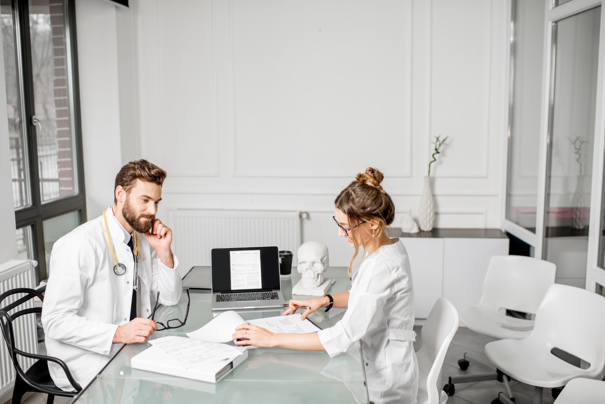 A male and female doctor in white coats sitting across from each other at a glass desk, reviewing printed documents with a laptop and anatomical skull model nearby.